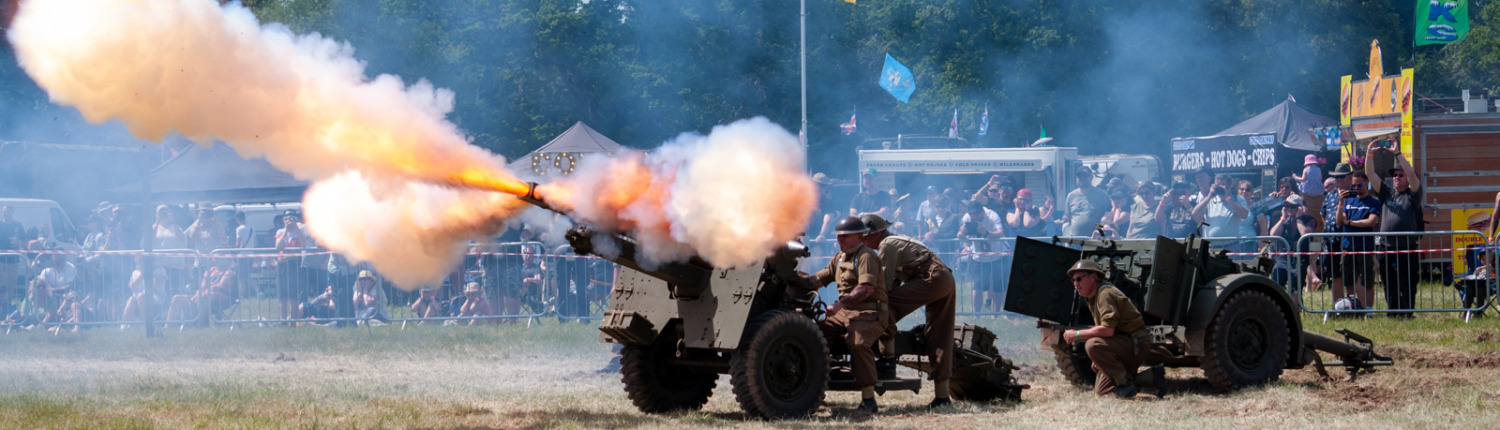 A soldier crew operates a howitzer, firing it surrounded by a crowd. Large flames erupt from the muzzle as the cannon is discharged.